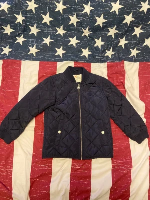 Mörkblå quiltad bomberjacka Holly & White - Quiltad bomberjacka i mörkblått från Holly & White. Jackan har ribbad krage, dragkedja framtill och två fickor med silverfärgade knappar. Materialet är syntet och jackan har långärmade ärmar. Perfekt för dig som gillar stilren och sportig look. Storlek 116. Jackan är i ganska så bra skick men det finns tecken på användning. 