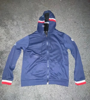 Blå Moncler hoodie med dragkedja - Snygg marinblå hoodie från Moncler med hel dragkedja och huva. Tröjan har röda och vita ränder på huvan och ärmsluten samt Moncler-logga på ena ärmen. Perfekt för dig som gillar streetwear och vill ha något stilrent men ändå sportigt.