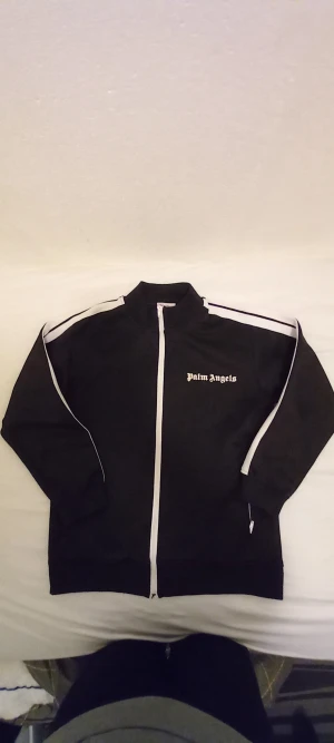 Svart track jacket från Palm Angels - Palm Angels svart track jacket med vita ränder längs ärmarna och vit dragkedja. Klassisk sportig look med logotyp på bröstet. Tillverkad i bomull med lite elastan för skön passform. Långärmad och med ståkrage.