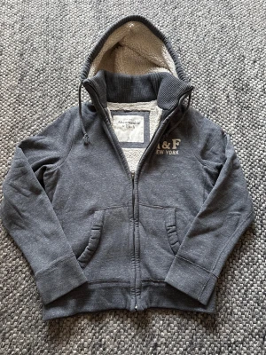 Grå teddycardigan Abercrombie & Fitch - Mysig grå teddyjacka från Abercrombie & Fitch med vit fluffig teddyfoder på insidan och huva. Jackan har dragkedja, ribbade muddar och broderad A&F New York-logga på bröstet. Perfekt för kyliga dagar och riktigt skön att bära. Väldigt fint skick!