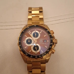 TAG Heuer Formula 1 guldarmbandsklocka - Säljer en TAG Heuer Formula 1 Calibre 16 kronograf med guldigt armband och boett. Klockan har en rund urtavla med svarta och vita detaljer, datumvisning och tachymeter på ringen. Robust design i metall med snygga tryckknappar på sidan. Nytt batteri behövs 