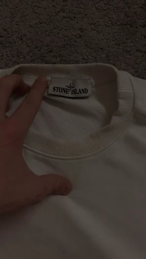 Vit långärmad Stone Island sweatshirt - Vit långärmad sweatshirt från Stone Island med klassisk rund hals och ribbade muddar. Tröjan har den ikoniska Stone Island-patchen på vänster ärm och är tillverkad i mjuk bomull. Perfekt för dig som gillar clean och stilren streetwear.