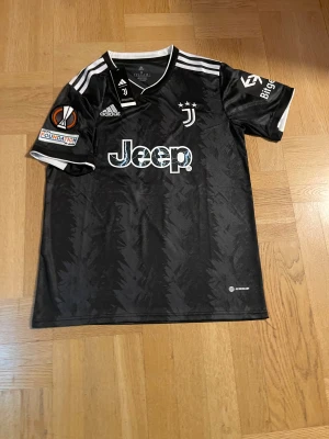 Juventus svart matchtröja Adidas - Juventus officiella svarta matchtröja från Adidas med vita detaljer och tryck. Tröjan har korta ärmar, UEFA Foundation-märke på ärmen och Jeep-logga på bröstet. Tillverkad i lätt och ventilerande polyester med subtilt mönster i tyget.