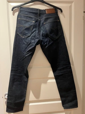 Mörkblå jeans från Tiger of Sweden - Säljer ett par snygga mörkblå jeans från Tiger of Sweden i klassisk femficksmodell. Jeansen har raka ben, kontrastsömmar och är tillverkade i Italien. Materialet är robust denim med en stilren look som passar perfekt till streetwear.