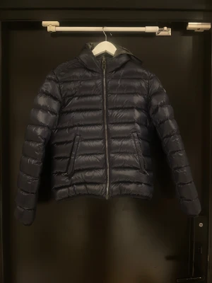 Marin blå Colmar pufferjacka med huva - Snygg svart pufferjacka från Colmar med glansig finish och huva. Jackan har två fickor med dragkedja och Colmar-logga på ärmen. Perfekt för kalla dagar. 