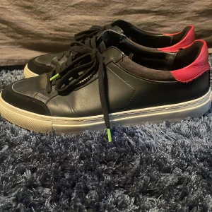 Svarta sneakers från Axel Arigato - Snygga svarta sneakers från Axel Arigato med klassisk vit sula och cool röd detalj på hälen. Skorna är i skinn och har svarta snören med små gröna detaljer. Perfekt för dig som gillar stilrena men ändå unika sneakers.