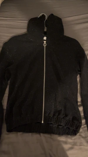 Svart glittrig bomberjacka från Lindex - Svart bomberjacka från Lindex med glittriga detaljer över hela tyget. Jackan har huva, lång ärm och en snygg silverfärgad dragkedja med rund ring. Perfekt för dig som vill sticka ut med lite extra bling i vardagen.