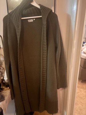 Brun stickad kofta från Basic - Säljer en olivgrön, lång och mysig stickad kofta från Basic. Koftan har bred ribbad kant längs öppningen och vid ärmsluten, samt en stor krage som ger extra chill vibe. Perfekt att slänga på sig när det är lite kyligt ute.