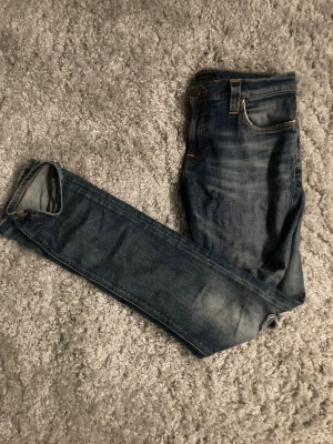 Blå jeans från Nudie Jeans - Säljer ett par klassiska blå jeans från Nudie Jeans med snyggt slitna detaljer och kontrastsömmar. Jeansen har fem fickor, normal passform och ikonisk broderad bakficka. Perfekta för dig som gillar en tidlös och avslappnad look.