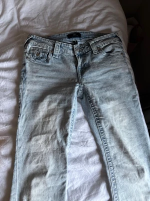 Ljusblå bootcut jeans från True Religion - Säljer ett par ljusblå jeans från True Religion med bootcut passform. Jeansen har klassiska fem fickor, snygga detaljerade bakfickor med broderi och silverfärgade knappar. Materialet är jeans i bomull och de har en avslappnad vibe med slitna detaljer.