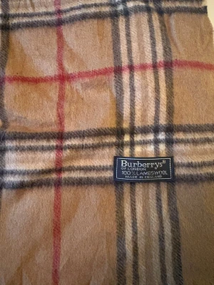 Rutig beige halsduk från Burberrys - Klassisk halsduk från Burberrys i beige med rutigt mönster i svart, vitt och rött. Gjord av 100% lammull och har snygga fransar i ändarna. Perfekt för att ge din outfit en lyxig touch och hålla dig varm under kyliga dagar.