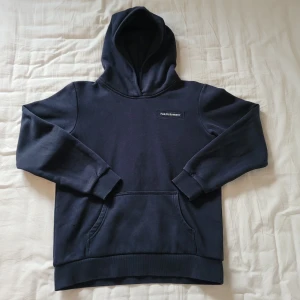 Mörkblå hoodie Peak Performance  - Snygg mörkblå hoodie från Peak Performance med klassisk logga på bröstet. Tröjan har huva, magficka och ribbade muddar vid ärmar och nederkant. Tillverkad i mjuk bomullsmix som är skön mot huden.