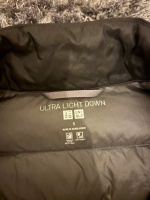 Svart dunväst från Uniqlo, storlek S - Svart, quiltad dunväst från Uniqlo Ultra Light Down. Västen har hög krage, dragkedja framtill och en clean, minimalistisk look. Perfekt för lager-på-lager och enkel att bära tack vare sin lätta känsla.