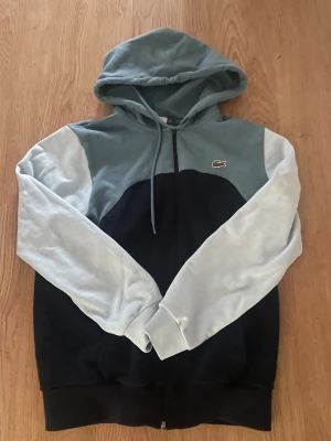 Lacoste hoodie i blå, ljus blå  - Snygg hoodie från Lacoste med färgblock i ljusblått, mörkblå och turkos . Tröjan har huva med snörning, dragkedja framtill och klassisk krokodillogga på bröstet. Perfekt för chill dagar och streetstyle.