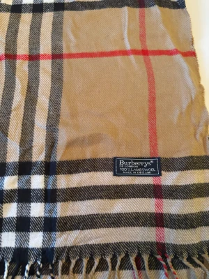Rutig beige halsduk från Burberrys - Klassisk halsduk från Burberrys i beige med rutigt mönster i svart, vitt och rött. Gjord av 100% lammull och har fransar i kanterna. Perfekt för att ge din outfit en snygg och tidlös touch.