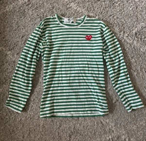 Randig långärmad tröja Comme des Garçons - Säljer min brorsas Comme des garçons tröja. Grön och vit randig långärmad tröja från Comme des Garçons Play, med det ikoniska röda hjärtat broderat på bröstet. Tröjan är i mjuk bomull och har en klassisk rund hals. Perfekt för dig som gillar streetstyle och coola detaljer. Han kunde ha den när han var runt 170 cm