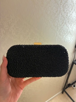 Svart glittrig kuvertväska med kedja - Snygg svart kuvertväska med glittriga pärlor över hela ytan och guldfärgade metalldetaljer köpt från Glitter. Passar perfekt till middagar och bal. Använt 1 gång och prislapp finns fortfarande kvar.