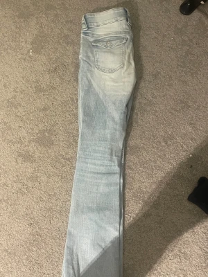 Bootcut ljusa jeans från H&M - Bootcut jeans från H&M i ljusblå tvätt med låg midja och justerbar midja. Klassisk femficksmodell med snygga detaljer som dubbla knappar och ficklock med knapp bak. Perfekt för dig som gillar retrovibbar och relaxed stil. Jag har vanligt vis S-M passar bra på mig
