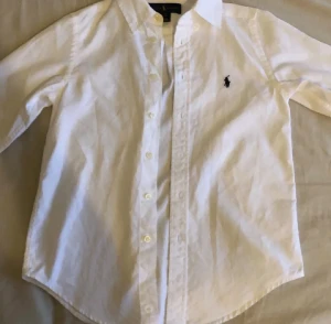 Vit skjorta från Ralph Lauren - Klassisk vit skjorta från Ralph Lauren med svart broderad logga på bröstet. Skjortan har lång ärm, knappar framtill och snygg krage. Passar dig som är runt 165. Pris kan diskuteras 