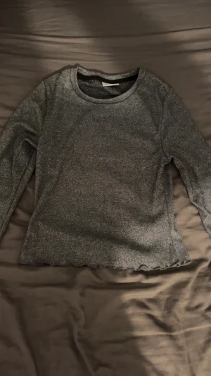 Glittrig långärmad tröja Kappahl - Snygg mörkgrå långärmad tröja från Kappahl med glittrigt material. Rund halsringning och vågig kant nedtill ger en cool touch. Perfekt för dig som gillar lite extra shimmer i vardagen.