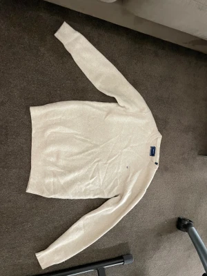 Beige stickad tröja från GANT - Snygg beige stickad tröja från GANT i superfin lammull. Klassisk rund hals och ribbade muddar vid ärmslut och nederkant. Diskret GANT-logo med broderad sköld på bröstet. Perfekt för dig som gillar stilrena och mjuka plagg.