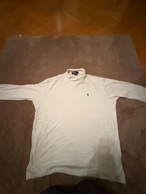 Vit polotröja från Polo Ralph Lauren - Bra julklapp!!! Vit polotröja från Polo Ralph Lauren med klassisk svart broderad logga på bröstet. Tröjan har lång ärm och hög krage som kan vikas ner. Mjuk och skön i bomull, perfekt för dig som gillar stilrena och tidlösa plagg.  (🔥om det syns några fleckar så är det sånt som går att tvätta 🧽 🔥)