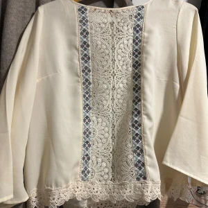 Vit blus med spets och broderi - Säljer en vit blus med lång ärm och vackra spetsdetaljer framtill samt längs nederkanten. Blusen har två smala blå broderade band som ramar in spetsen och ger en unik look. Perfekt för dig som gillar romantiska och boho-vibbar.