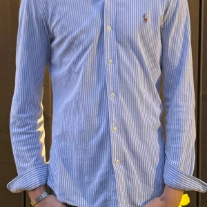 Blårandig skjorta från Ralph Lauren - Klassisk blårandig skjorta från Ralph Lauren med button down-krage och broderad logga på bröstet. Skjortan har lång ärm, vita knappar och är tillverkad i mjuk bomull. Perfekt för en clean och stilren look.