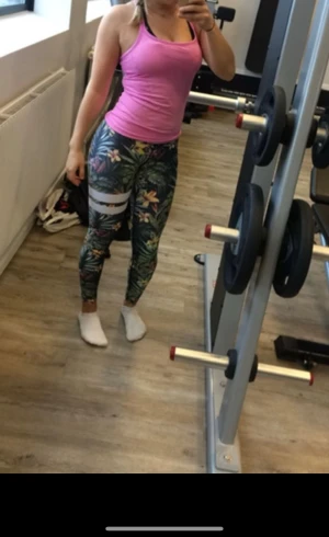 Blommiga leggings från Stronger - Snygga träningstights med tropiskt blommönster i grönt, gult, rosa och vitt. De har två vita ränder på ena benet och bred vit linning. Perfekta för gymmet eller träning, sitter tight och är gjorda i stretchigt syntetmaterial. Litet litet hål vid midjan, se sista bild 