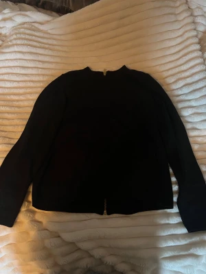 Svart tröja med dragkedja i bak från Stockh LM - Svart tröja från Stockh LM i storlek S. Tröjan har en hel dragkedja i guld längs hela baksidan och är ribbad vid halsringning och ärmslut. Långärmad och i mjukt stickat material, perfekt för lager-på-lager under kyliga dagar.