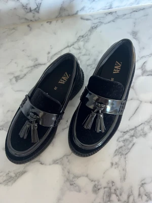 Svarta loafers med tofsar från Zara - Snygga svarta loafers från Zara med glansiga detaljer och tofsar framtill. Skorna har en rund tå och platt sula, kombinerar mocka och syntetmaterial för en cool look. Perfekta för dig som gillar klassisk stil med en modern twist.