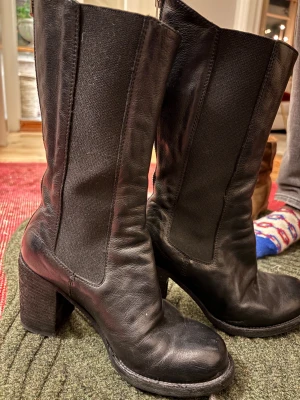 Svarta stövletter med klack från officine creative - Säljer ett par svarta stövletter i skinn från officine creative. De har chunky klack, rund tå och dragkedja längs hela baksidan. Snygga detaljer med elastiska paneler på sidorna och text på yttersulan. Perfekta för dig som gillar edgy och stilren look.