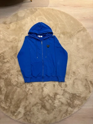  zip hoodie från Comme des Garçons Play - Säljer en blå zip hoodie från Comme des Garçons Play med dragkedja och huva. Tröjan har ett svart hjärta med ögon på bröstet och ett stort tryck med samma motiv på ryggen. Tillverkad i mjuk bomull och har snörning vid huvan samt praktiska fickor framtill. Kom eventuellt för frågor 🧐pris kan diskuteras 💯 .