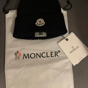 Svart ribbad Moncler mössa - Svart ribbstickad mössa från Moncler med klassisk logotyp framtill. Mössan har uppvikt kant och är tillverkad i mjukt material som håller dig varm. Perfekt accessoar för kyliga dagar och snygg till streetwear eller vinterjacka.
