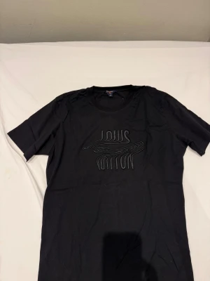 ÄKTA Svart Louis Vuitton t-shirt  - Passar storlek M Svart t-shirt från Louis Vuitton med coolt broderat logotypmönster på bröstet och en orange patch med Louis Vuitton Paris på ryggen. Rund hals och korta ärmar, tillverkad i mjuk bomull. Made in Italy.