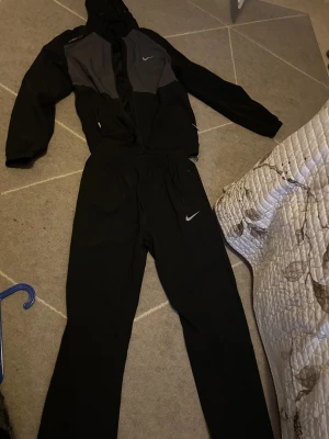 Nike svart och grå tracksuit - Nike travksuit  med svart och grå zip-hoodie och matchande svarta byxor. Jackan har huva, dragkedja och snörning i midjan, samt Nike-logga på bröstet. Byxorna har elastisk midja med snörning och vit Nike-logga på höften. Perfekt för sport och chill.