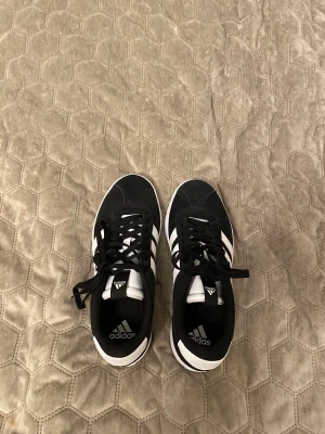 Svarta Adidas sneakers med vit plattform - Säljer ett par svarta Adidas sneakers med klassiska vita ränder på sidorna och vit plattformsula. Skorna har snörning och rund tå, samt en snygg kontrast mellan svart mocka och vita detaljer. Perfekta för dig som gillar stilrena och sportiga vibes.
