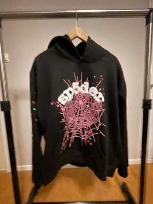 Svart Sp5der hoodie - Svart hoodie från Sp5der i storlek S med stor rosa spindelnätsprint och vita 3D-bokstäver på bröstet. Hoodien har huva och långa ärmar med små stjärnor i rött och blått som detaljer. Perfekt för dig som gillar streetwear med unik design. Storlek M passar också L.
