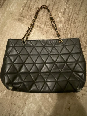 Svart quiltad axelväska med kedja - Svart axelväska med quiltat triangelmönster och guldfärgad kedja som axelrem. Väskan är tillverkad i skinnimitation och har en klassisk rektangulär form. Perfekt för dig som gillar stilrena detaljer och vill ha en väska med lite edge. Vissa defekter som ni ser, därav pris🌸