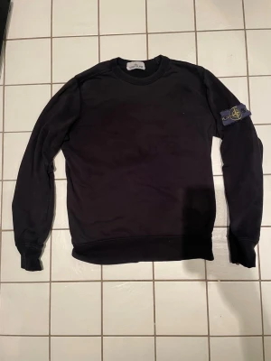 Svart sweatshirt från Stone Island - Svart långärmad sweatshirt från Stone Island med klassisk rund hals och ikonisk patch på vänster ärm. Tröjan har en clean look och är tillverkad i mjuk bomull som är skön mot huden. Perfekt för dig som gillar streetwear och vill ha något stilrent. Storlek S passar storlek XS.