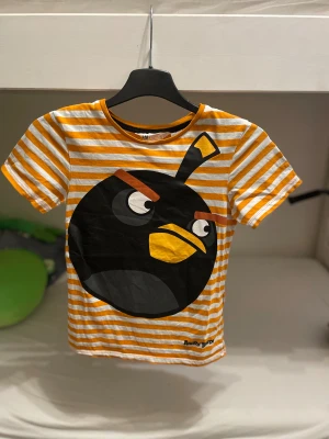 Randig Angry Birds t-shirt H&M - Cool t-shirt från H&M med Angry Birds-tryck framtill. T-shirten är vit och orange med ränder och har en stor svart Angry Birds-figur på bröstet. Klassisk passform och korta armar. Tillverkad i mjuk bomull, perfekt för dig som gillar färg och gaming. Barn till pojkar, storlek 122/128