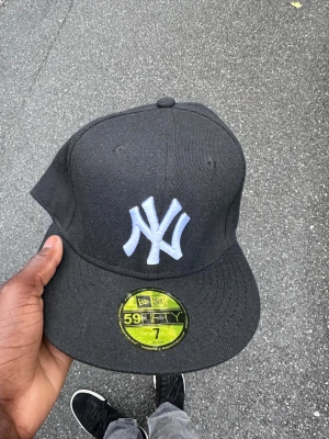 Svart New Era 59FIFTY NY keps - Säljer en svart New Era 59FIFTY keps med klassiskt New York Yankees broderat i vitt framtill. Kepsen har en rak skärm och är tillverkad i ett slitstarkt bomullsmaterial. Perfekt för dig som gillar streetwear och sportig stil.
