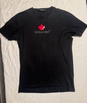 Svart Dsquared2 t-shirt med lönnblad - Svart t-shirt från Dsquared2 i storlek M. Framsidan har ett tryck med rött lönnblad och Dsquared2-logga i grått. Klassisk rund hals och korta ärmar. Materialet är mjuk bomull som känns skön mot huden. Perfekt för dig som gillar stilrena designer med en twist.