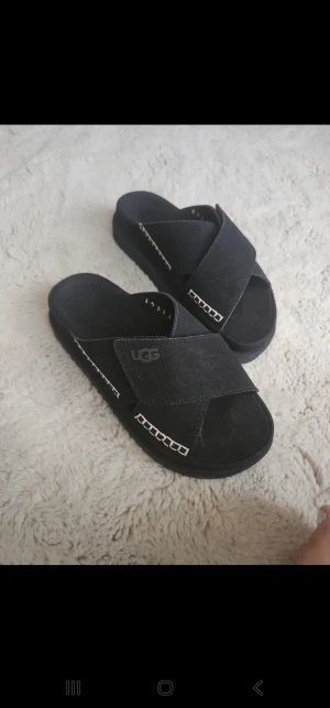 Svarta sandaler  med korsade band - Svarta sandaler med breda korsade band i mocka och dekorativa vita sömmar längs kanterna. Platt sula med mönstrad undersida för extra grepp. UGG-logga på sidan och öppen tå för chill vibe.