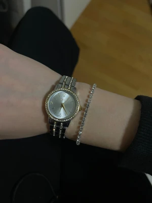 Regal silver och guld klocka med stenar - Snygg klocka från Regal med rund urtavla i silver och gulddetaljer. Boetten är dekorerad med glittriga stenar runt kanten och armbandet är i silver och guld med infällda stenar. Klockan har en klassisk och stilren look som drar blickarna till sig.