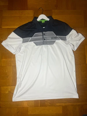 Boss  - Snygg vit och mörkblå henley från Boss med stilrena ränder över bröstet och klassisk krage. Skjortan har korta ärmar, knappar framtill och logga på bröstet. Materialet är mjukt och lätt polyester, perfekt för en fräsch look.