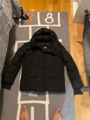 Svart wyndham jacka från Canada Goose  - Säljer en svart dunjacka från Canada Goose med klassisk logga på ärmen. Jackan har huva, långa ärmar och knappar framtill. Den är quiltad och har en clean, stilren look som funkar när det är kallt ute. Perfekt för dig som vill ha en varm jacka med street vibe.