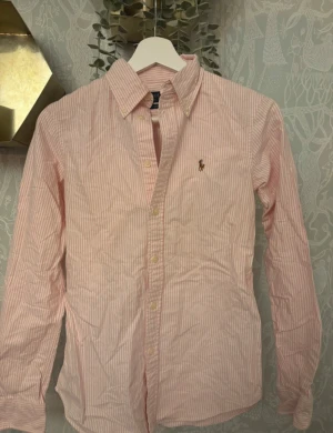 Rosa randig skjorta från Polo Ralph Lauren - Snygg rosa och vit randig skjorta från Polo Ralph Lauren i modellen Custom Fit. Skjortan har klassisk krage, broderad logga på bröstet och knappar framtill. Tillverkad i mjuk bomull och har långärmad passform som sitter riktigt nice.