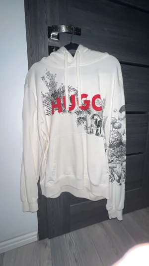 Vit hoodie från HUGO med print - Säljer en vit hoodie från HUGO med stor röd logga och detaljerat svart print med motiv av palmer och skateboards på framsidan, ärmen och ryggen. Hoodien har snörning i huvan och en stor magficka. Perfekt för dig som gillar streetstyle och grafiska tryck. 