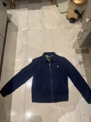 Mörkblå vindjacka från Polo Ralph Lauren - Snygg mörkblå vindjacka från Polo Ralph Lauren med klassisk krage, dragkedja framtill och broderad logga på bröstet. Jackan har rutigt foder i grönt, blått och gult samt två fickor på sidorna. Perfekt för dig som gillar stilren och tidlös design. Lite sol blekt men inget stort